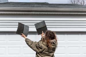 Young woman female homeowner standing in front of house garage in coat jacket during winter storm holding two roof tile shingles inspecting damage