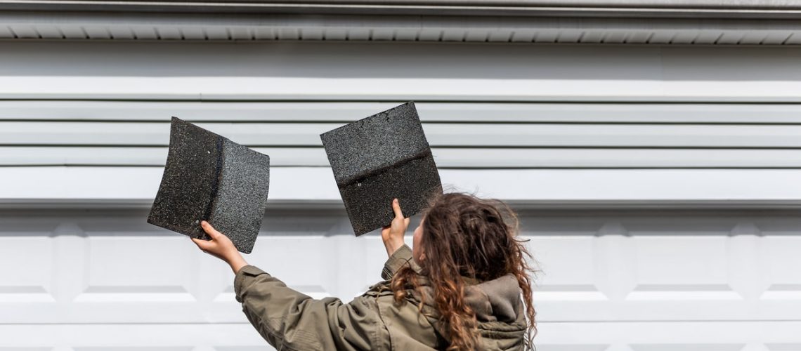 Young woman female homeowner standing in front of house garage in coat jacket during winter storm holding two roof tile shingles inspecting damage